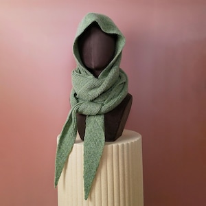 May include: A sage green knitted hood and scarf set displayed on a mannequin head. The scarf is wrapped around the neck and drapes down with pointed ends. The hood is up, showcasing the texture of the knit fabric. The mannequin is on a white column.