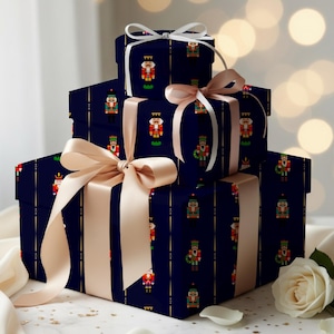 Könnte beinhalten: Drei gestapelte quadratische Geschenkboxen, eingewickelt in dunkelblaues Papier mit einem sich wiederholenden Nussknacker-Muster. Die Boxen sind mit breiten, champagnerfarbenen Satinbändern und Schleifen versehen. Eine weiße Rose liegt daneben.