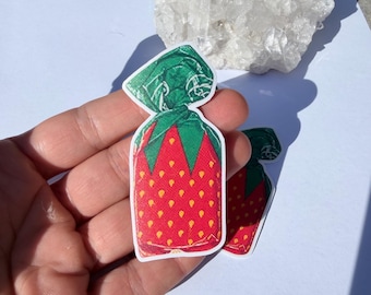 May include: A die-cut sticker shaped like a strawberry candy wrapped in green foil. The candy features a bright red body with yellow seed-like patterns and a green twisted top. The sticker has a white border and a matte finish, showing a graphic design.