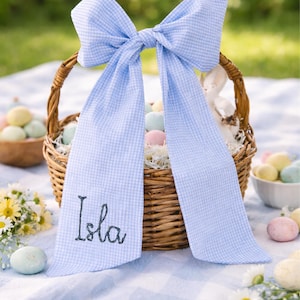 May include: An Easter basket made of wicker, adorned with a large blue and white gingham bow. The bow features the name "Isla" embroidered on one tail. The basket contains decorative eggs and rests on a blue and white checked blanket.