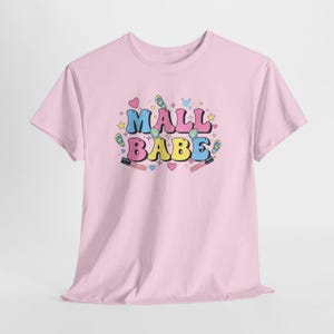 Può includere: T-shirt rosa chiaro con la scritta "MALL BABE" in lettere colorate. Il design include cuori, stelle e tubetti di rossetto. Un capo casual per tutti i giorni.