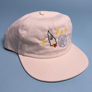 Puede incluir: Gorra de béisbol beige claro con visera curva. La gorra presenta diseños bordados de un cohete, estrellas y un planeta en hilo amarillo, negro, rojo y azul. La gorra tiene un botón en la parte superior y está sobre un fondo azul claro.
