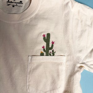 Puede incluir: Una camiseta blanca con un bolsillo que presenta un cactus bordado con flores rosas. La camiseta tiene el texto "Animal Instincts Apparel" en la parte superior.
