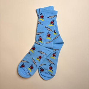 May include: A pair of light blue socks with a repeating pattern of gray narwhals and red and yellow flames. The socks are made of a soft material and are designed to fit a standard foot size.