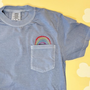 May include: A light blue short-sleeve t-shirt with a chest pocket. An embroidered rainbow design in red, orange, yellow, green, blue, and purple is visible peeking out from the top of the pocket. The inner neck label reads Comfort Colors S P CH.