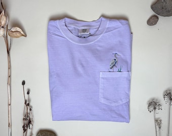 May include: A light purple short-sleeve t-shirt with a crew neckline and a single chest pocket. The pocket features an embroidered graphic of a blue heron standing in tall grass. The interior neck label displays the text Comfort Colors and size L.