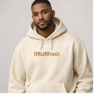 May include: Cream-colored hoodie with the word "NAIJAfresh" printed in a brown font across the chest. The hoodie has a drawstring hood and a front pocket. The person in the image is wearing the hoodie.