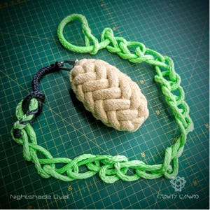 Pode incluir: Um objeto ovalado bege tecido à mão, com uma corrente de corda verde e um laço de corda preta e verde. A corrente e o laço são conectados ao oval com um fecho de metal. O texto "Nightshade Oval" é visível.