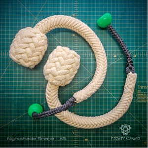 Könnte beinhalten: Ein beiges Seilspielzeug mit zwei geflochtenen Knoten und einem gebogenen Design. Das Spielzeug hat einen dunkelgrauen geflochtenen Abschnitt und grüne Kunststoff-Endstücke. Der Text "Nightshade Snake - X6" ist sichtbar.