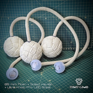 May include: Three off-white, woven fire poi with curved rope handles and LED grips. The poi heads are approximately 8.5 cm in diameter. The text at the bottom reads "85 mm Titan + Static Kevlar + Ultra Knob Pro LED Grips."