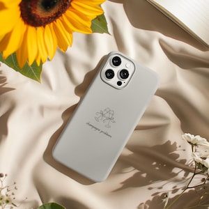 May include: A light gray phone case with the words "champagne problems" and a champagne glass design. The phone case is lying on a beige fabric, with a sunflower and white flowers in the background.