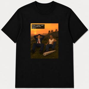 Puede incluir: Una camiseta negra de manga corta con un estampado gráfico de dos personas sentadas en una colina cubierta de hierba al atardecer. Uno lleva traje y corbata, el otro una camiseta blanca sin mangas. Un objeto grande enrollado yace en el césped. Texto: Dawg, I swear I told you to grab the lighter!