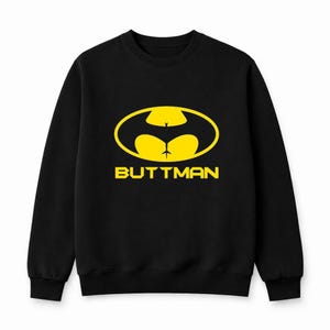 May include: Black sweatshirt with a yellow graphic of a bat-shaped design with a buttock-like shape in the center. The word "BUTTMAN" is printed in yellow below the graphic.