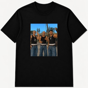 Könnte beinhalten: Ein schwarzes T-Shirt mit kurzen Ärmeln und einem rechteckigen Grafikdruck auf der Brust. Der Druck zeigt drei Figuren, die im Freien stehen und jeweils ein schwarzes Unterhemd, eine blaue Jeans und ein weißes Unterhemd tragen.