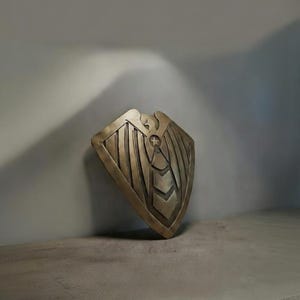 May include: A bronze-colored shield with a detailed design, featuring vertical lines and a central emblem. The shield is slightly angled, resting against a gray wall, and positioned on a concrete shelf.