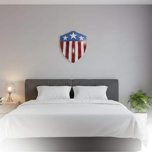 Op de afbeelding: Een slaapkamer met een bed met witte lakens en kussens. Boven het bed hangt een schildvormige wanddecoratie in rood, wit en blauw, met sterren en strepen. Een nachtkastje met een lamp en een kleine plant.