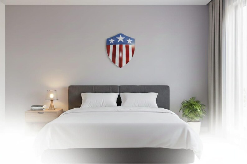 Op de afbeelding: Een slaapkamer met een bed met witte lakens en kussens. Boven het bed hangt een schildvormige wanddecoratie in rood, wit en blauw, met sterren en strepen. Een nachtkastje met een lamp en een kleine plant.
