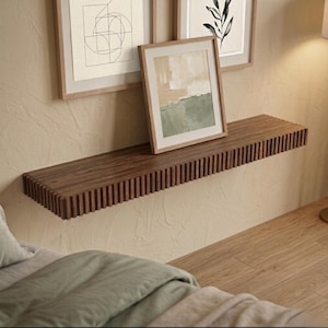 Op de afbeelding: Een donkerbruin zwevend houten plank met een groefvormig randontwerp. De plank is bevestigd aan een lichtbeige muur, met ingelijste kunstwerken erboven. Een ingelijst landschapsschilderij rust op de plank. Het algemene thema is moderne woondecoratie.