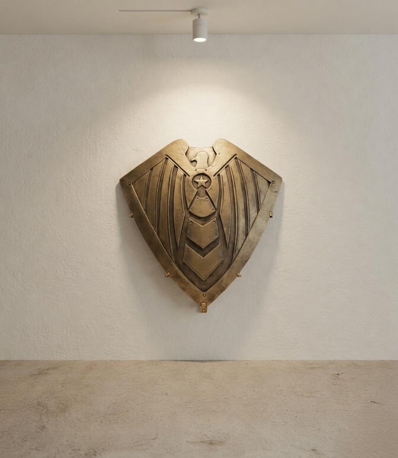 May include: A bronze-colored shield with an eagle emblem and star design, mounted on a white wall. The shield has a central star and wing-like patterns. The shield is a decorative wall hanging.