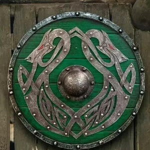 May include: Circular, green Viking shield with a detailed silver dragon design. The shield features a central metal boss and is trimmed with silver studs. The dragon artwork has red accents. The shield is mounted on a wooden background.