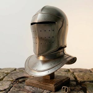 Puede incluir: Un casco de caballero medieval de metal, con visera y protector de cuello. El casco se exhibe sobre un soporte de madera. El metal tiene una apariencia desgastada, lo que sugiere edad o uso. El diseño incluye pequeños orificios de ventilación.