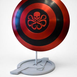 May include: A red and black circular shield with a skull and octopus design in the center. The shield is mounted on a gray stand with the Avengers logo. The shield is a prop or collectible.