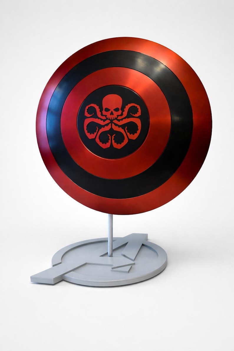 May include: A red and black circular shield with a skull and octopus design in the center. The shield is mounted on a gray stand with the Avengers logo. The shield is a prop or collectible.