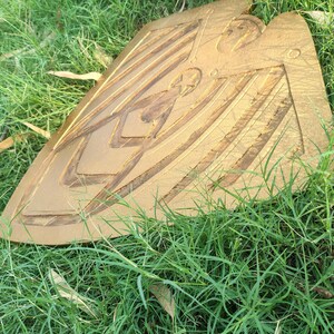 May include: A golden-colored, shield-shaped object with carved geometric designs. The shield is resting on a bed of green grass. The shield has a star and other shapes carved into it.