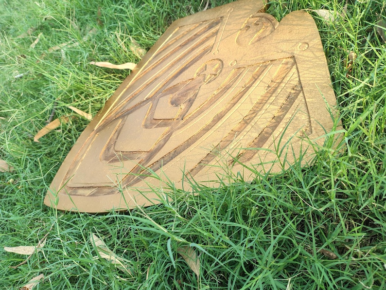 May include: A golden-colored, shield-shaped object with carved geometric designs. The shield is resting on a bed of green grass. The shield has a star and other shapes carved into it.