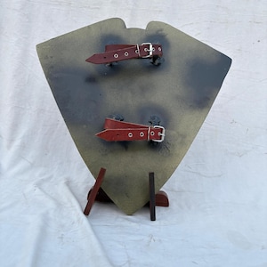 May include: A shield-shaped object with a camouflage pattern in shades of green, brown, and gray. It features two red leather straps with silver buckles. The shield is supported by a wooden stand.