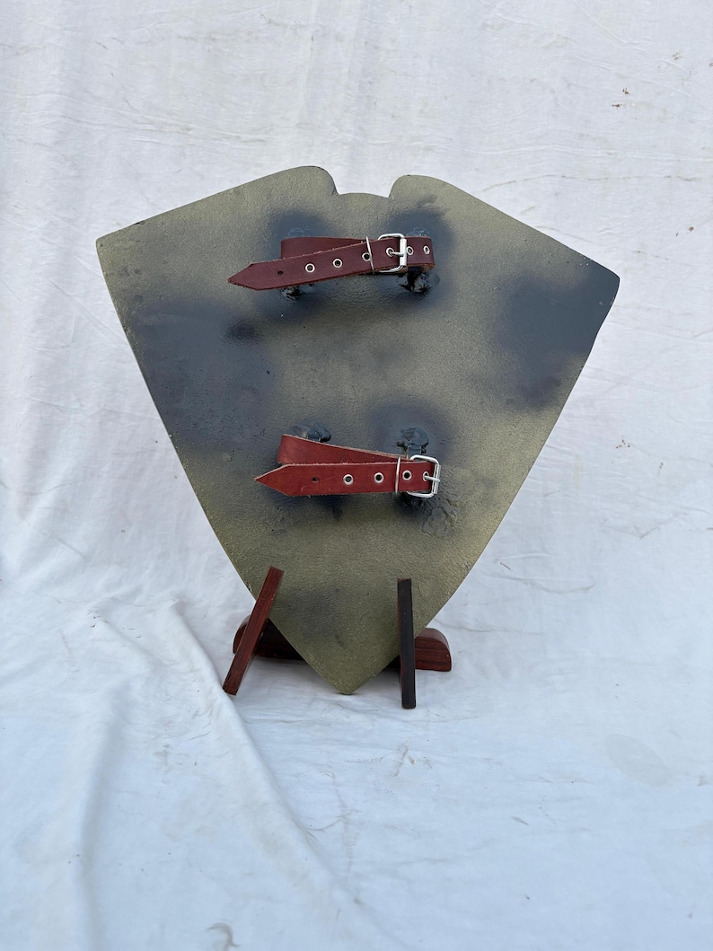 May include: A shield-shaped object with a camouflage pattern in shades of green, brown, and gray. It features two red leather straps with silver buckles. The shield is supported by a wooden stand.