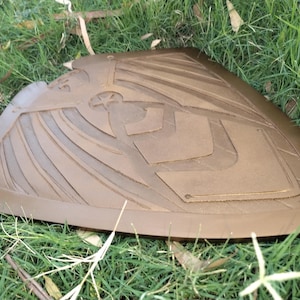 May include: A brown, shield-shaped object with an embossed design of an eagle and geometric shapes. The shield is resting on a bed of green grass and fallen leaves. The shield appears to be made of leather or a similar material.