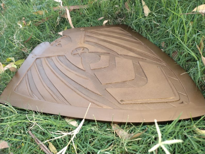 May include: A brown, shield-shaped object with an embossed design of an eagle and geometric shapes. The shield is resting on a bed of green grass and fallen leaves. The shield appears to be made of leather or a similar material.