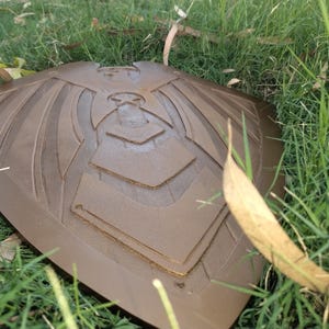 May include: A bronze-colored shield with an embossed design of a stylized bird or eagle, resting on a bed of green grass. The shield has a curved shape with detailed lines and geometric patterns.