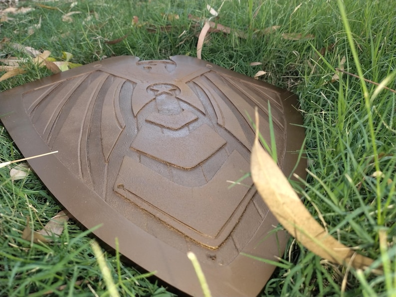 May include: A bronze-colored shield with an embossed design of a stylized bird or eagle, resting on a bed of green grass. The shield has a curved shape with detailed lines and geometric patterns.