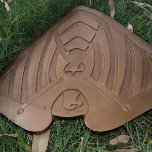 May include: A bronze-colored, shield-shaped object with intricate carved details. The design includes geometric shapes, a star, and curved lines. The object is resting on a bed of green grass.