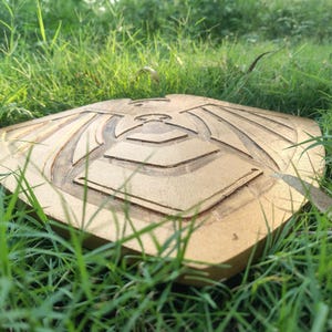 May include: A gold-colored, square-shaped wooden shield with an engraved design of a stylized bird or eagle. The shield is lying on a bed of green grass, with the sun shining on it.