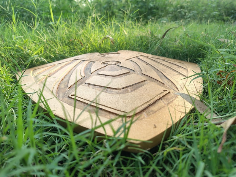 May include: A gold-colored, square-shaped wooden shield with an engraved design of a stylized bird or eagle. The shield is lying on a bed of green grass, with the sun shining on it.
