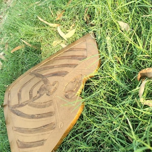 May include: A golden-brown, shield-shaped object with carved linear and curved designs, possibly a decorative piece or a part of a larger artwork. The object is resting on a bed of green grass, suggesting an outdoor setting.