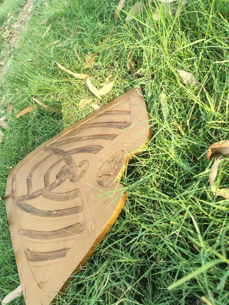 May include: A golden-brown, shield-shaped object with carved linear and curved designs, possibly a decorative piece or a part of a larger artwork. The object is resting on a bed of green grass, suggesting an outdoor setting.