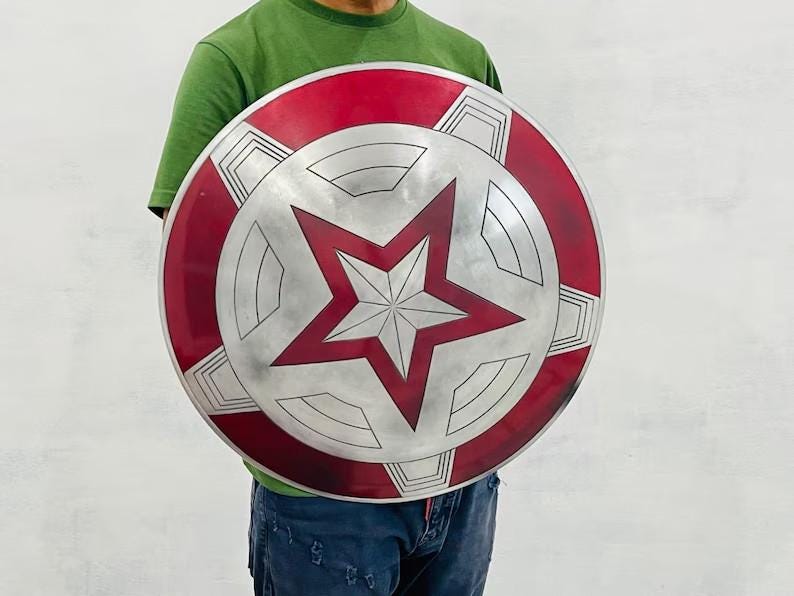 May include: A round shield with a red and silver design. The outer rim is red, with silver geometric shapes. The center features a large, red, five-pointed star on a silver background. A classic superhero accessory.