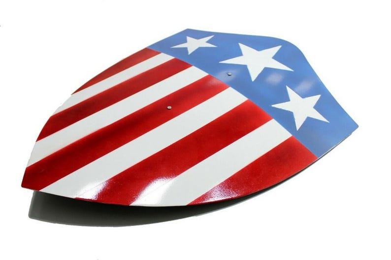 Op de afbeelding: Een schild met een patriottisch ontwerp, met rode en witte strepen en een blauw gedeelte met witte sterren. Het schild heeft een gebogen vorm en een glanzende afwerking, die doet denken aan het embleem van een superheld.