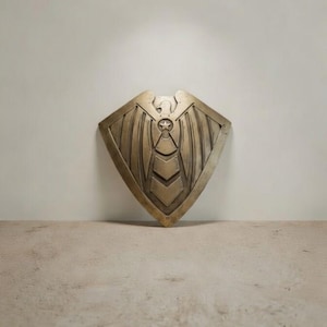 May include: A bronze-colored shield with an eagle and star design. The shield has a diamond shape with a central star and chevron pattern. The eagle is at the top of the shield, with wings spread. The shield is made of metal.