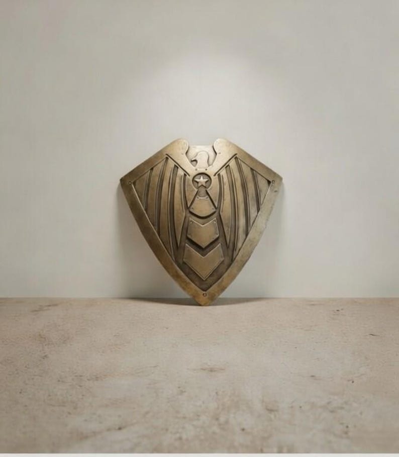 May include: A bronze-colored shield with an eagle and star design. The shield has a diamond shape with a central star and chevron pattern. The eagle is at the top of the shield, with wings spread. The shield is made of metal.