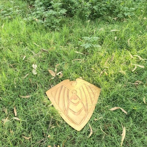 May include: A golden-colored, shield-shaped wooden carving with an eagle-like figure at the top and a central design. The carving is placed on a bed of green grass, with a natural outdoor setting.