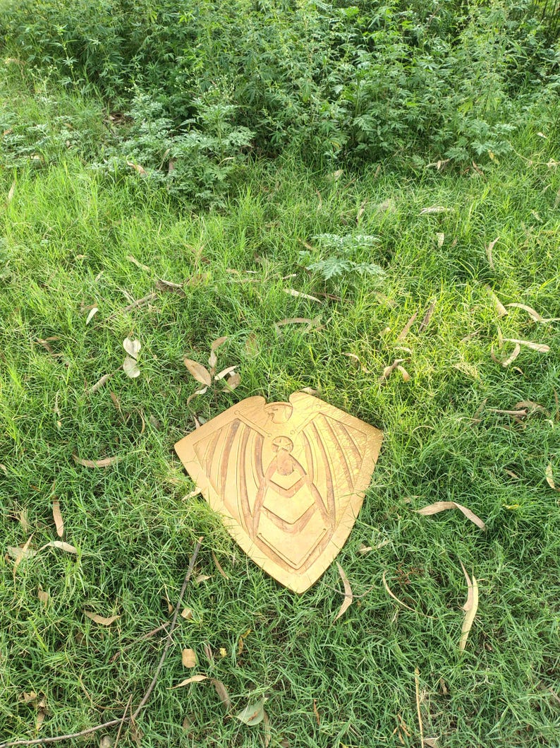 May include: A golden-colored, shield-shaped wooden carving with an eagle-like figure at the top and a central design. The carving is placed on a bed of green grass, with a natural outdoor setting.