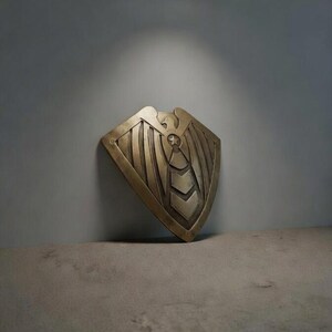 May include: A bronze-colored shield with an eagle design. The shield has a curved top and a series of vertical lines and chevron shapes. The shield is leaning against a gray wall.
