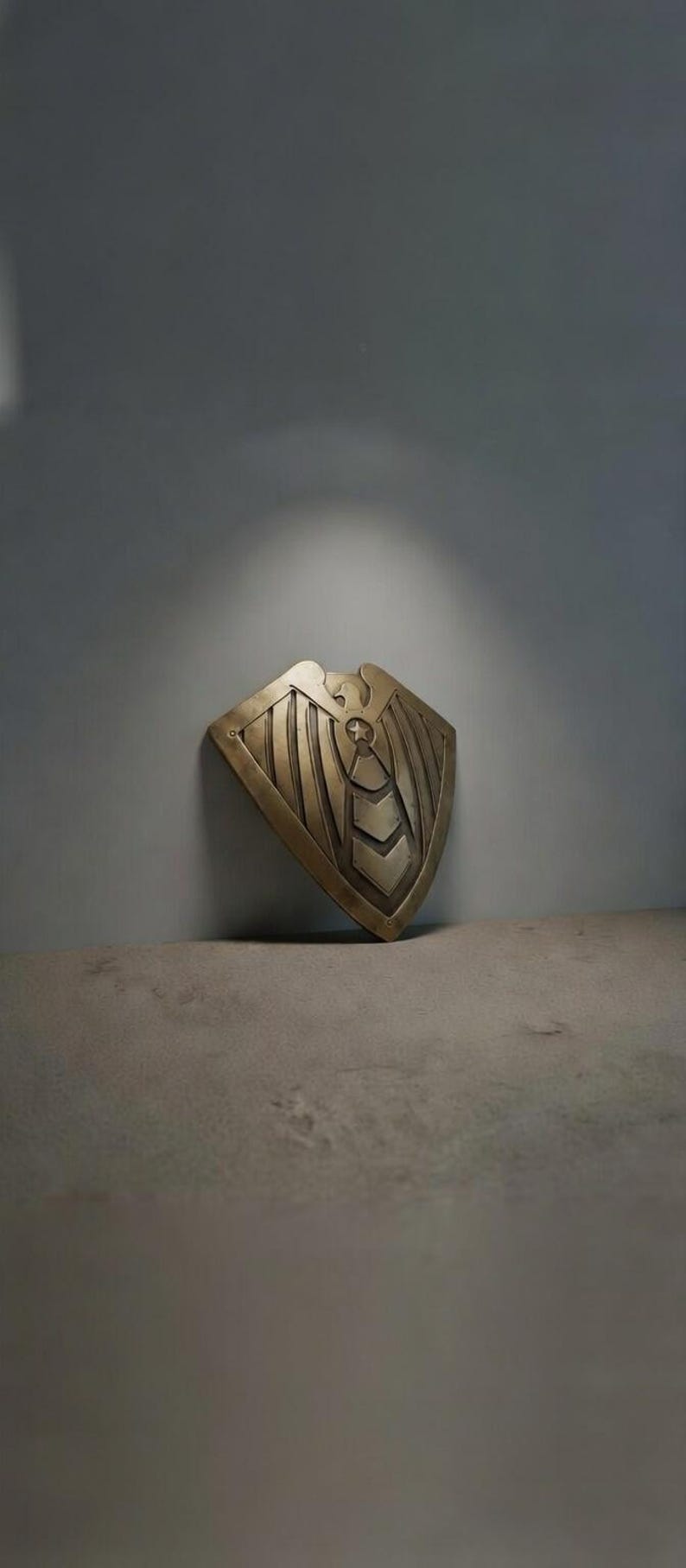 May include: A bronze-colored shield with an eagle design. The shield has a curved top and a series of vertical lines and chevron shapes. The shield is leaning against a gray wall.