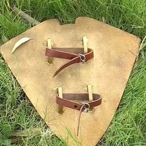 May include: A tan leather shield with two brown leather straps and brass rivets. The shield is diamond-shaped and rests on green grass.