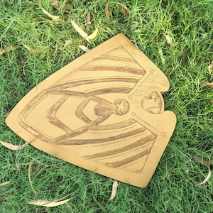 May include: A wooden shield with a star and geometric design, resting on green grass. The shield has a light brown color with darker brown etched details. The design includes a star, lines, and a figure.